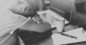 An exhausted woman sleeping on her laptop, her head in her hands, represents the looming threat of AI to writers.