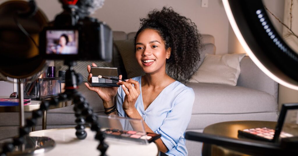 A woman holding a makeup mirror and a camera, leveraging tools and resources for her beauty routine blog content.