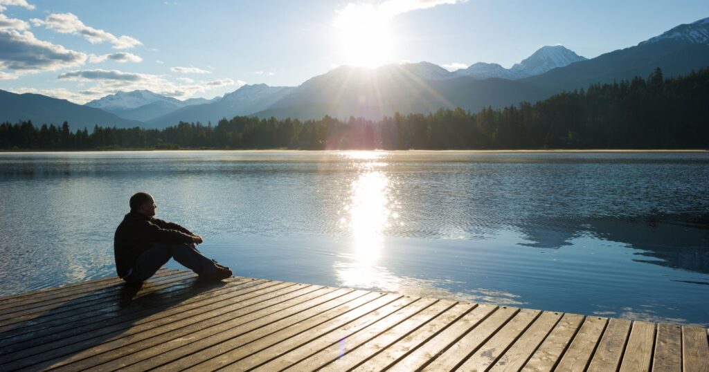 A man sitting on a dock, gazing at the sun as it sets. Reflecting on the moment, he adapts to the changing world.